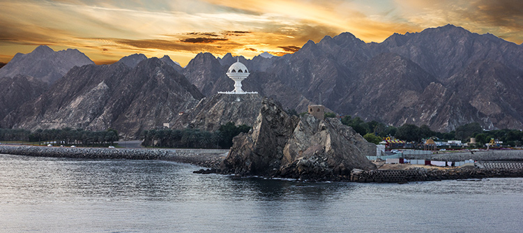 The sun sets behind the giant incense burner and dramatic rocky mountains on the shore of Muscat, Oman.